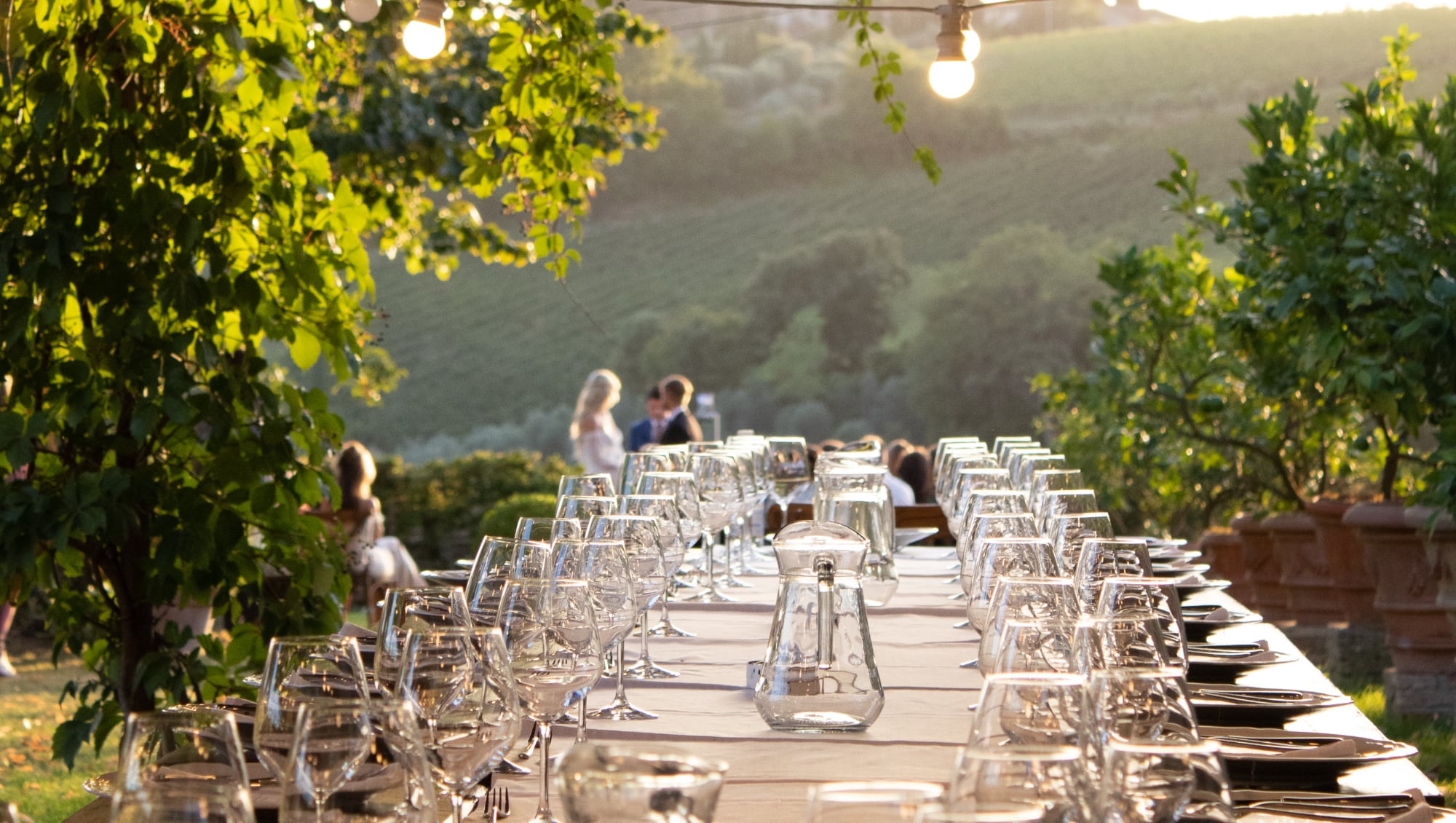 Long outdoor dinner table set with wine glasses overlooking Tuscan vineyard hills at sunset during a wedding reception