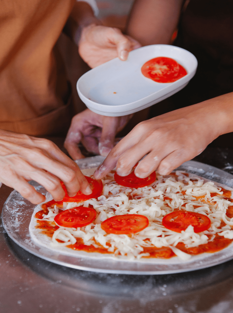 Hands topping a hand-stretched pizza with mozzarella and tomato slices during a pizza-making class